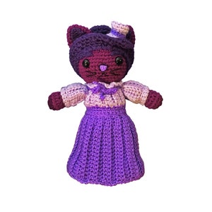 May include: A purple and pink crocheted cat doll wearing a hat and a dress. The doll has black eyes and a black nose.