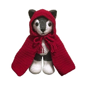 May include: A crocheted wolf plush toy wearing a red hooded cape. The wolf has a grey face and white body with black eyes and a black nose.