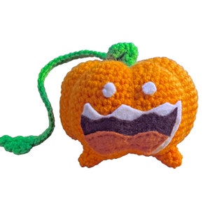 May include: A crocheted orange pumpkin with a green stem and a stitched-on white and brown face. The pumpkin has a wide, toothy grin.