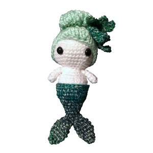 May include: A crocheted mermaid doll with a green and white tail and a green bun. The doll has large black eyes and a white body.