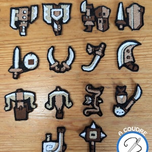 Könnte beinhalten: Eine Sammlung gestickter Aufnäher mit verschiedenen Waffendesigns in Braun-, Beige- und Weißtönen. Die Aufnäher sind auf einer Holzoberfläche angeordnet und zeigen detaillierte Stickereien und eine Vielzahl von Formen, darunter Schwerter, Äxte und Bögen.