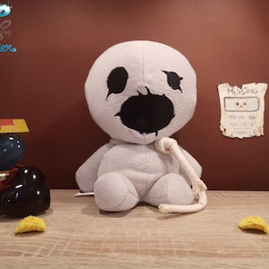 Op de afbeelding: Een lichtgrijs pluche karakter met grote zwarte ogen en mond, en een wit touw om zijn nek. Een "Missing" poster staat op de muur. Een schatkist en andere decoratieve items zijn ook aanwezig. Het karakter is een pluche speelgoed.