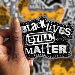 May include: A black and white sticker with the text "BLACK LIVES STILL MATTER" in a distressed font. The sticker has a stitched border and is held against a background of similar stickers.