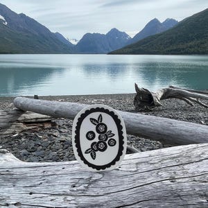 Pode incluir: Um prato oval de cerâmica branca com uma borda preta recortada. O prato apresenta uma ilustração preta de mirtilos com folhas. O fundo mostra um lago e montanhas.