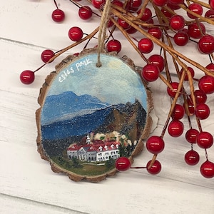 May include: A wooden ornament with a painted scene of a white building with red roofs and a mountain range in the background. The ornament is painted in shades of blue, green, white, and red. The text "Estes Park" is written on the ornament in white paint.