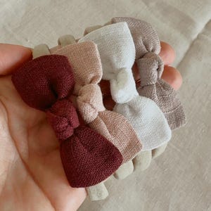 May include: A collection of fabric hair ties in various colors, including burgundy, pink, white, and beige. The hair ties are arranged in a circular pattern, showcasing their bow-like design. The background is a neutral, light-colored fabric.