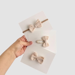 May include: Three baby hair accessories: a headband, a barrette, and a clip. Each features a bow made of tan and white checkered fabric. The headband has a thin brown elastic band. The barrette and clip have metal hardware.