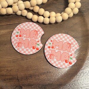 May include: Two round car coasters with a pink and white checkered pattern. The word "MAMA" is printed in pink bubble letters with cherry accents. The coasters are on a wooden surface.