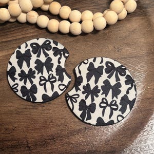 May include: Two round car coasters with a black bow pattern on a white background. The coasters have a black border and a small notch for easy removal. The coasters are on a wooden surface with a string of wooden beads.
