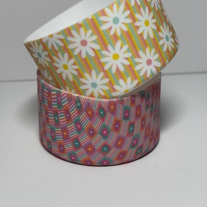 May include: Two stacked cylindrical bowls. The top bowl features a white daisy pattern over vertical stripes in yellow, pink, and teal. The bottom bowl displays a checkered pattern of small pink and teal flowers against a background of thin stripes.