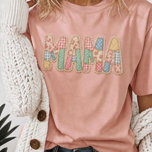 Peut inclure: Un t-shirt rose clair à manches courtes avec le mot MAMA écrit en grandes lettres style bloc. Chaque lettre est remplie d'un patchwork de motifs floraux, vichy et à pois dans des tons pastel, bordé d'une bordure festonnée décorative.