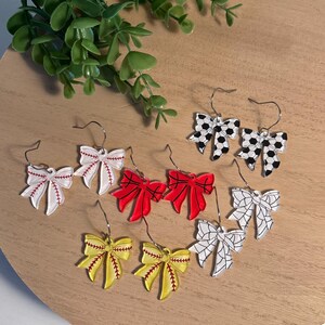 May include: A collection of bow-shaped earrings in various colors and designs. The earrings include baseball, softball, and soccer ball patterns. The earrings are displayed on a wooden surface with green foliage in the background.