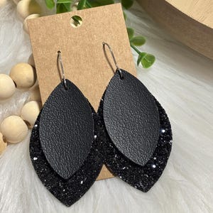 May include: A pair of black teardrop earrings. The earrings have a top layer of textured black material and a bottom layer of black glitter. The earrings are attached to silver-colored fish hook ear wires. The earrings are displayed on a brown card.