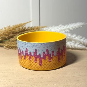 May include: A cylindrical silicone boot for a tumbler featuring a yellow waffle cone pattern at the base, topped with a layer of pink ice cream dripping down, and a light blue layer with small sprinkles covering the top rim of the accessory.