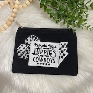 May include: Black zippered pouch with embroidered patches. The patches include dice, a lightning bolt, and a rectangular patch with the text "Raising Hell with the Hippies and the Cowboys."