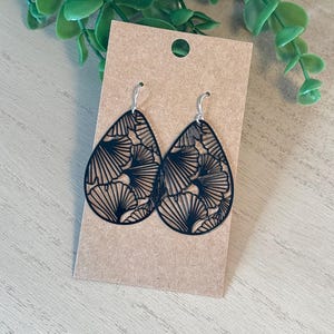 May include: Black teardrop earrings with a detailed, openwork design. The earrings are displayed on a brown card with a green plant in the background. The earrings have a silver hook.