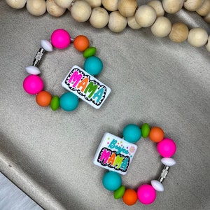 May include: Two colorful bracelets with the word "MAMA" on a white rectangular charm. The bracelets feature bright pink, orange, green, and teal beads. The charms have a black and white cow print border.