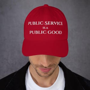 May include: A red baseball cap with white embroidered text that reads "Public Service is a Public Good".