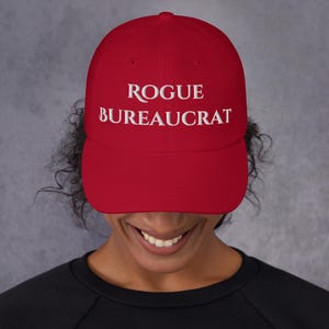 May include: A red baseball cap with white embroidered text that reads "ROGUE BUREAUCRAT".