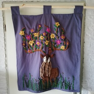 May include: A purple fabric wall hanging with a deer silhouette embroidered with brown felt and surrounded by a floral design with yellow and purple felt flowers.