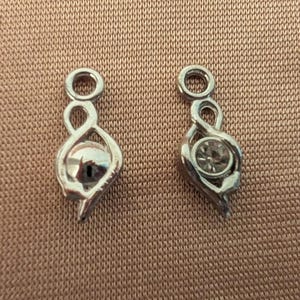 May include: Two silver-toned charms with a teardrop design and a small loop at the top. One charm has a clear, reflective stone, and the other has a faceted, smoky-colored stone. The charms are displayed on a textured, tan surface.