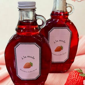 Puede incluir: Dos botellas de vidrio llenas de líquido rojo, con una etiqueta que dice "à la mode Strawberry Coffee Syrup". Cada botella tiene una tapa plateada y un asa. Una fresa fresca descansa sobre una superficie rosa.
