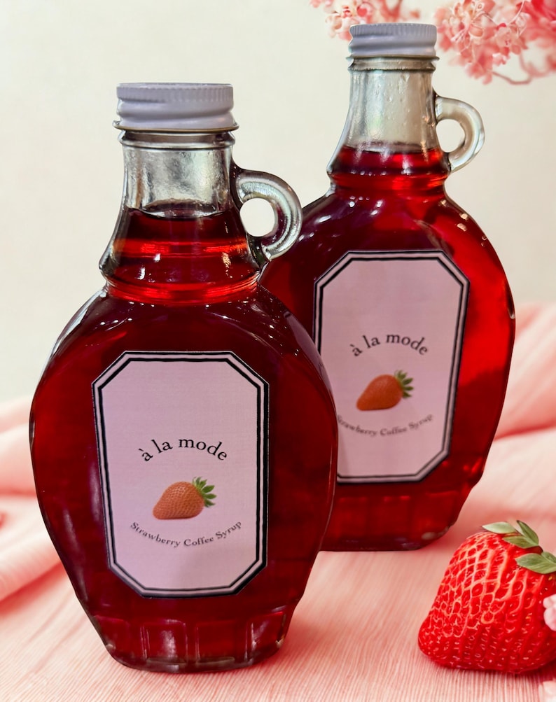 May include: Two glass bottles filled with red liquid, featuring a label that reads "&agrave; la mode Strawberry Coffee Syrup." Each bottle has a silver cap and a handle. A fresh strawberry rests on the pink surface.