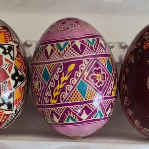 May include: Three decorated eggs arranged in a row. The left egg features a complex geometric star pattern in red, orange, and black. The center egg displays purple, teal, and yellow floral and diamond motifs. The right egg shows a yellow rooster.