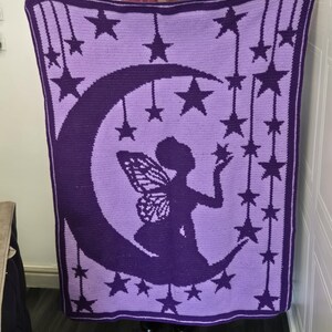 May include: A rectangular crochet blanket featuring a dark purple silhouette of a fairy with wings sitting on a crescent moon against a light purple background. The design includes numerous dark purple stars hanging from vertical lines.