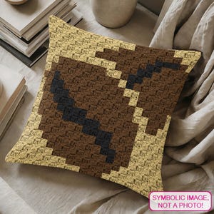 May include: A square crochet cushion featuring a pixelated coffee bean design. The pattern uses light beige, medium brown, and dark charcoal yarn to create the graphic. The texture is consistent with a granny square stitch, forming a blocky image.