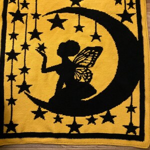 May include: A rectangular crochet blanket featuring a yellow background with a black border. The centre displays a black silhouette of a fairy with wings sitting on a crescent moon, surrounded by black stars hanging from strings.