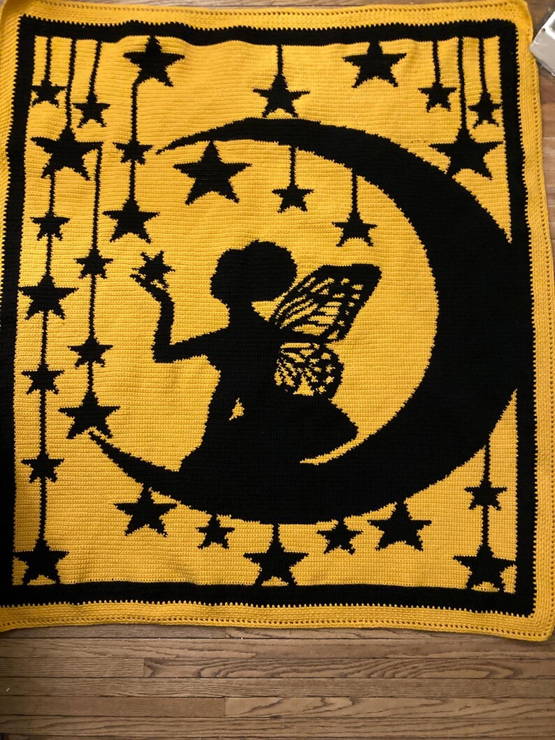 May include: A rectangular crochet blanket featuring a yellow background with a black border. The centre displays a black silhouette of a fairy with wings sitting on a crescent moon, surrounded by black stars hanging from strings.