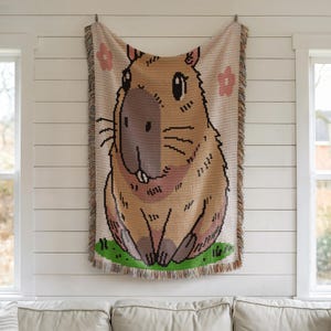 Puede incluir: Un tapiz de pared tejido con un capibara de dibujos animados con la nariz rosa y el pelaje marr&oacute;n. El capibara est&aacute; rodeado de flores rosas y hierba verde.