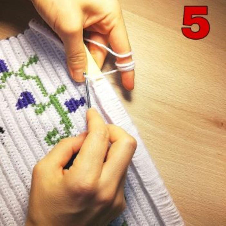 Pu&ograve; includere: Primo piano di una mano che usa un uncinetto per intrecciare filato bianco in un tessuto all'uncinetto bianco con un motivo floreale. Il tessuto viene lavorato su un telaio di legno.