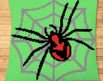 Op de afbeelding: Een groen gehaakt kussen met een zwarte spin op een grijs web. De spin heeft een rood lichaam en zwarte poten. De tekst "SYMBOLISCH BEELD, GEEN FOTO!" staat onderaan het kussen.