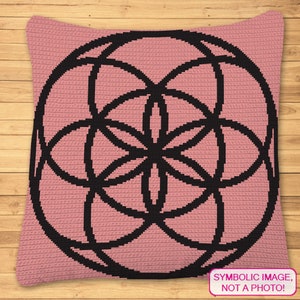 May include: A pink crocheted pillow with a black geometric design in the center. The design is a circle with overlapping circles inside, creating a flower-like pattern. The text "SYMBOLIC IMAGE, NOT A PHOTO!" is in a white box at the bottom of the pillow.