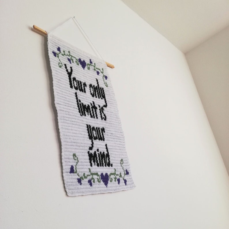 Pu&ograve; includere: Arazzo da parete in crochet bianco con il testo "Your only limit is your mind." in lettere nere. L'arazzo &egrave; decorato con cuori viola e viti verdi.