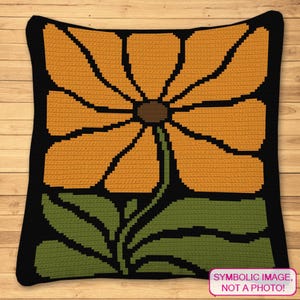Op de afbeelding: Een vierkant, gehaakt kussen met een groot, gestileerd oranje bloemdesign. De bloem heeft een bruin hart en staat tegen een zwarte achtergrond. Groene bladeren en stelen zijn zichtbaar. Het kussen is een decoratief woonaccessoire.