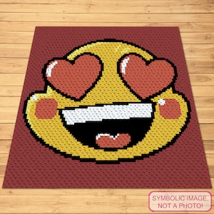 Puede incluir: Un emoji amarillo con corazones por ojos y una amplia sonrisa. El emoji está tejido a crochet con hilo rojo, blanco y amarillo.