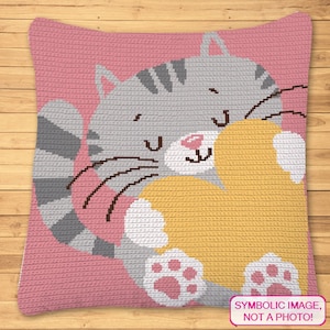May include: A gray and white crocheted cat pillow holding a yellow heart-shaped pillow. The cat has a pink nose and paws. The background is pink.