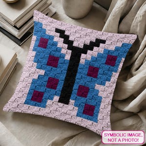Könnte beinhalten: Ein quadratisches, gehäkeltes Kissen mit einem pixeligen Schmetterlingsdesign. Der Schmetterling hat blaue Flügel mit dunkelvioletten Quadraten, einen schwarzen Körper und ist vor einem hellrosa Hintergrund platziert. Das Kissen liegt auf einer neutralfarbenen Oberfläche. Text im Bild: SYMBOLISCHES BILD, KEIN FOTO!