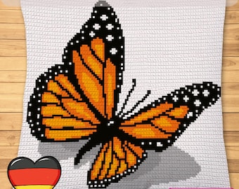 Op de afbeelding: Een wit gehaakt kussen met een groot oranje, zwart en wit vlinderontwerp. Het kussen heeft bovenaan de tekst "DEUTSCH HÄKELANLEITUNG" en links onderaan een hart met de Duitse vlag. De rechter benedenhoek heeft de tekst "SYMBOLIC IMAGE, NOT A PHOTO!"