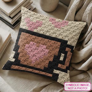 May include: A square crochet cushion cover featuring a pixelated coffee mug design in brown and black yarn. The mug contains a pink heart pattern, set against a cream-coloured background with additional pink heart accents at the top corners.