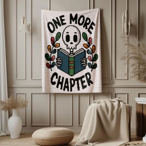 May include: A white crochet wall hanging featuring a pixelated skull reading a blue and green book. The text "ONE MORE CHAPTER" is stitched in black above and below the skull. Two colorful leafy branches frame the central design.