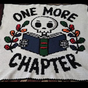 May include: A white crochet blanket featuring a central graphic of a skull reading a blue and green book. The text ONE MORE CHAPTER is stitched in black above and below the skull. Two leafy branches with red and green accents flank the book.