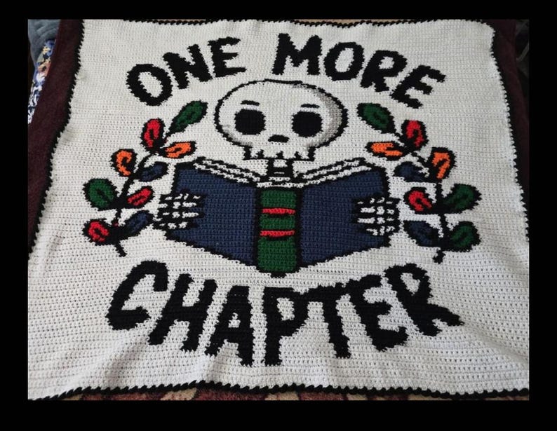 May include: A white crochet blanket featuring a central graphic of a skull reading a blue and green book. The text ONE MORE CHAPTER is stitched in black above and below the skull. Two leafy branches with red and green accents flank the book.