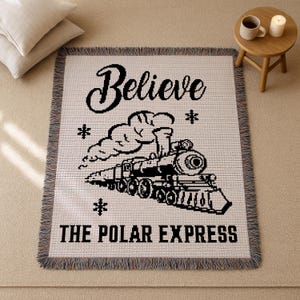 Puede incluir: Una manta tejida blanca con una ilustración negra de un tren y las palabras "Believe THE POLAR EXPRESS". La manta tiene un borde con flecos y está sobre una superficie beige. Cojines decorativos y una mesita con una taza y una vela están en el fondo.