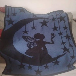 May include: A rectangular crochet blanket featuring a dark blue silhouette of a fairy sitting on a crescent moon. The design includes multiple black stars hanging from strings against a light blue background with a dark blue border.