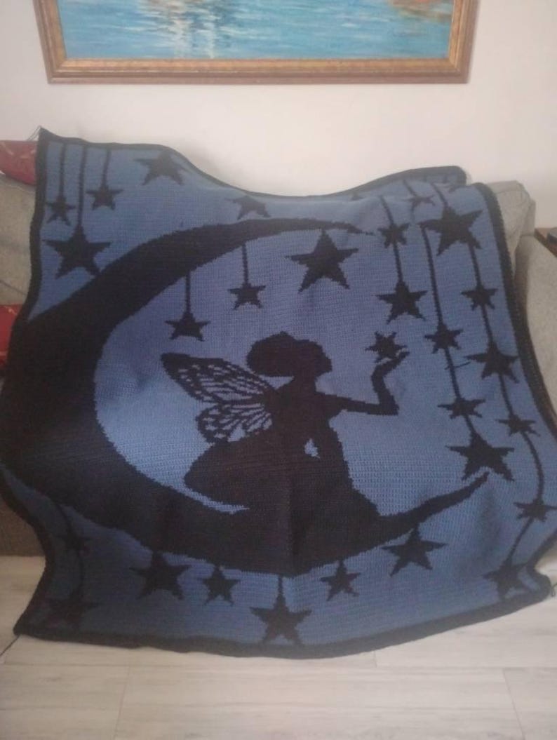 May include: A rectangular crochet blanket featuring a dark blue silhouette of a fairy sitting on a crescent moon. The design includes multiple black stars hanging from strings against a light blue background with a dark blue border.