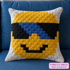 May include: A square crochet pillow featuring a yellow pixelated face with black sunglasses and a black smile. The design includes blue accents within the sunglasses and a white border. A pink rectangular label reads: SYMBOLIC IMAGE, NOT A PHOTO!